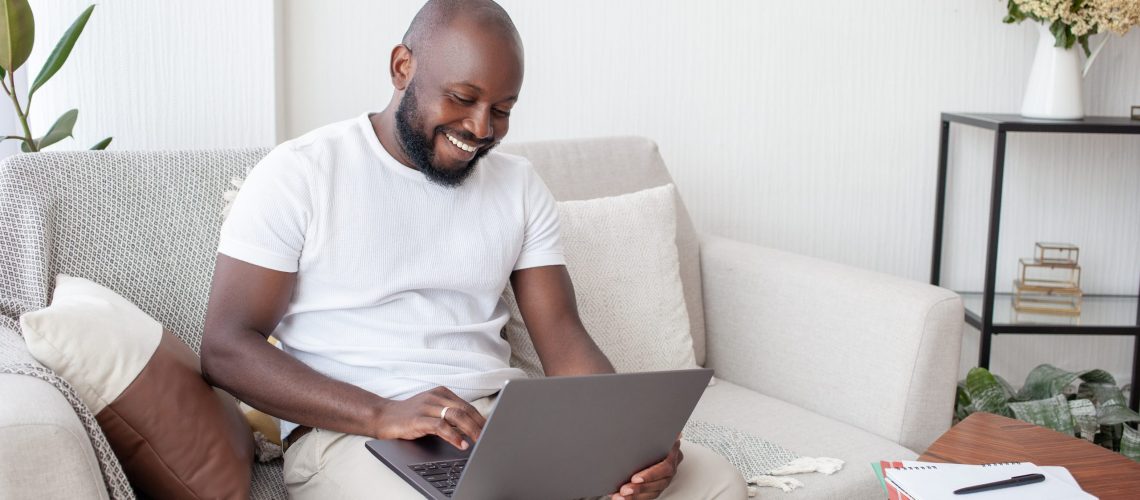 Afro-American man in a white T-shirt sitting on the sofa at home works remotely on a laptop, uses wireless Internet. People, technology and freelance concept.