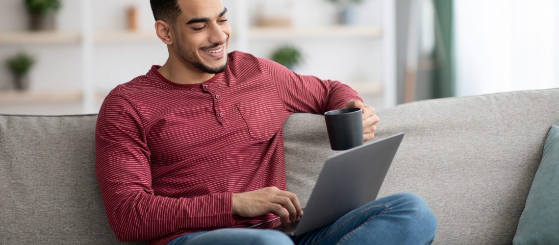 Smiling arab guy chatting with ladies online and drinking coffee, using dating website on laptop while resting on sofa at home. Happy muslim man watching movies or funny videos on notebook, copy space